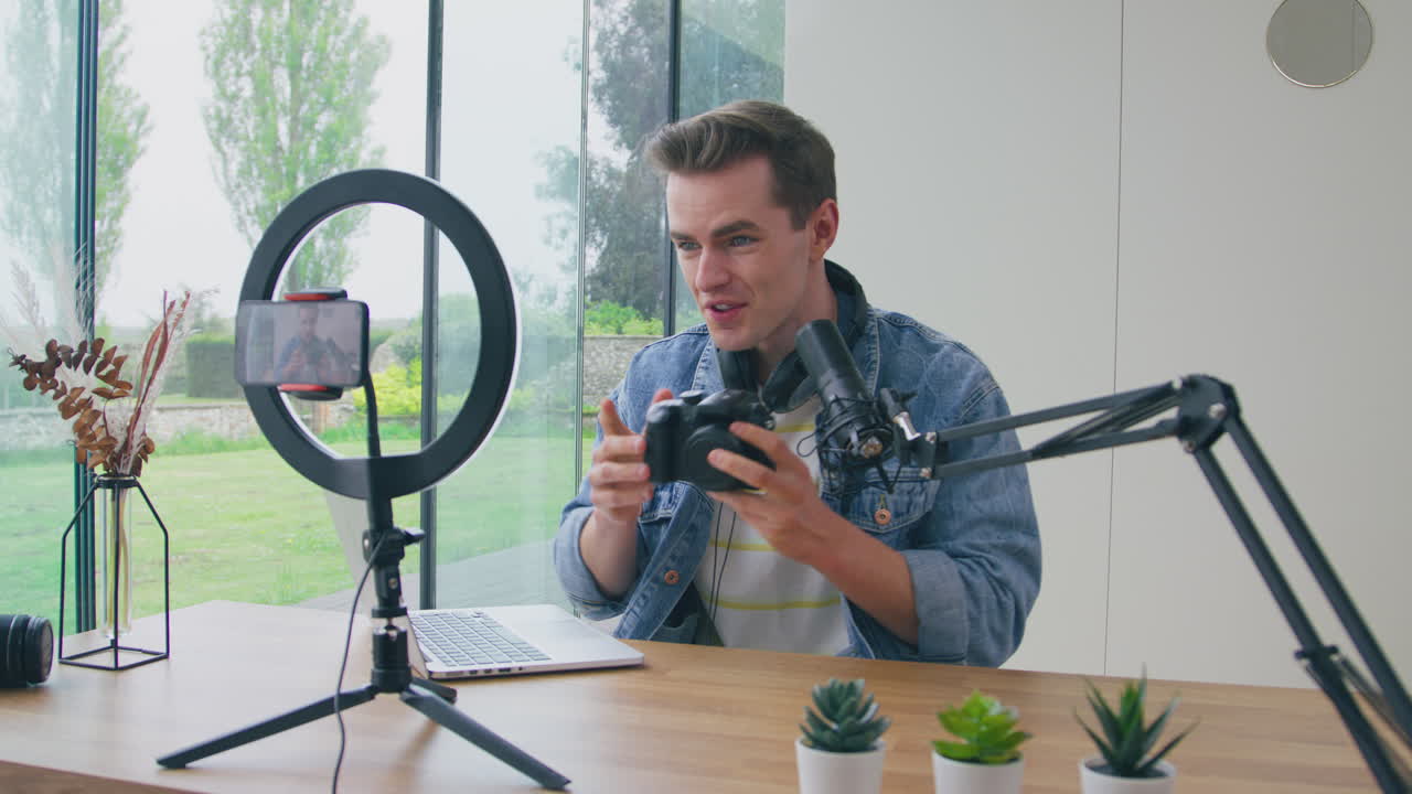 Mandlig vlogger live-streamer videoanmeldelse af kameraprodukt hjemmefra, optaget med mobiltelefon.