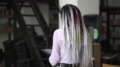 rare view of walking woman with long, black and white dreadlocks, looking on tablet in her hands. walking by modern, public library