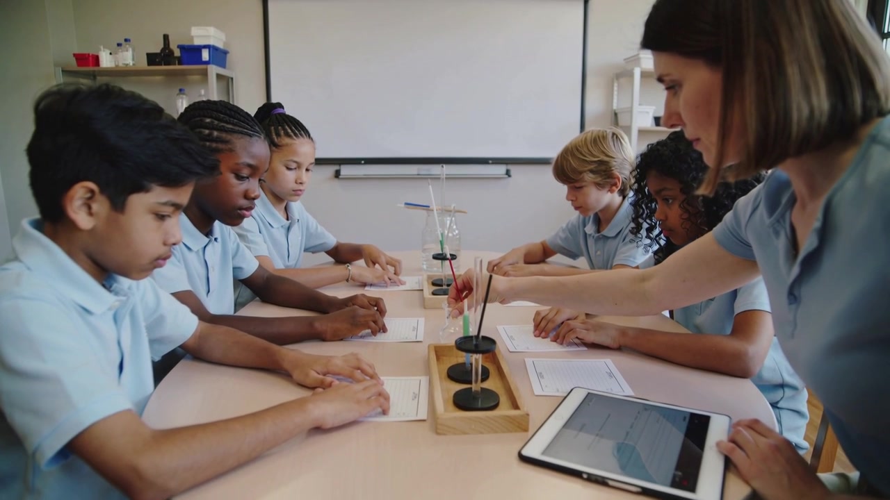 Reaching teacher guiding students in blue shirts at table, dipping brush into wells for art lesson