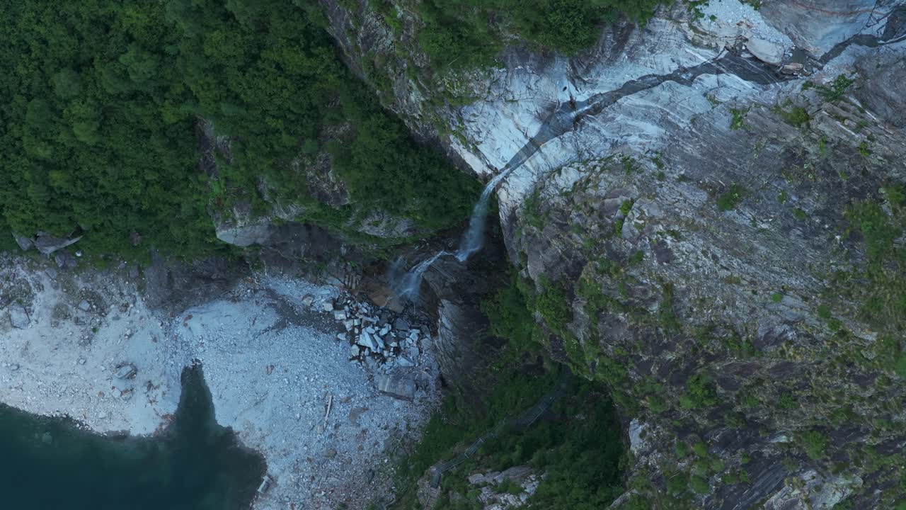 Top-down drone view of waterfall flowing over steep rocks into alpine Lake Antrona, surrounded by green forest, Italian Alps, Italy
