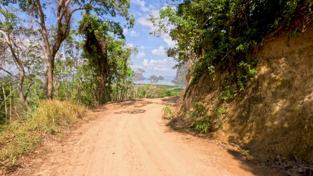 Camera glides along jungle dirt road, revealing panoramic limestone cliffs under bright daylight