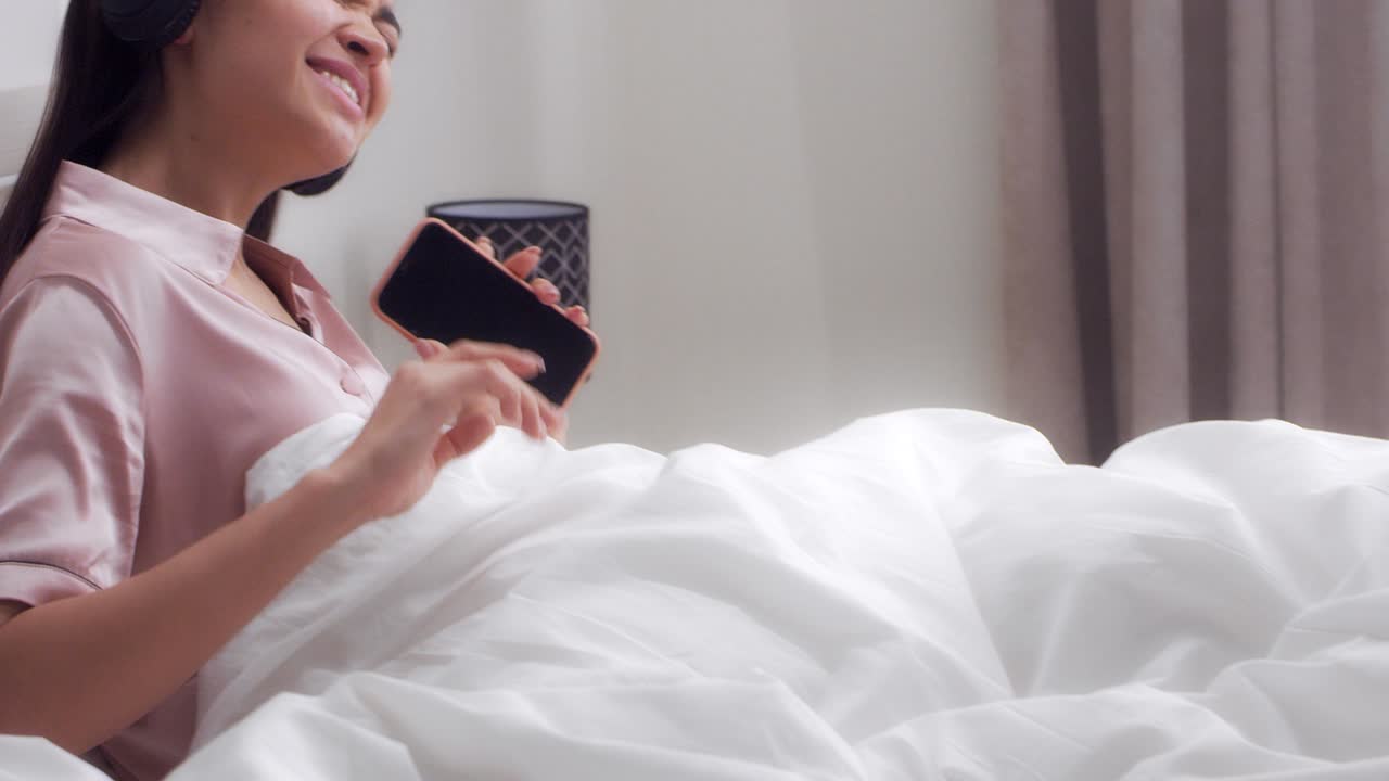 여자는 헤드폰을 통해 음악을 듣고 침대에 누워서 노래하고 춤을 춘다.