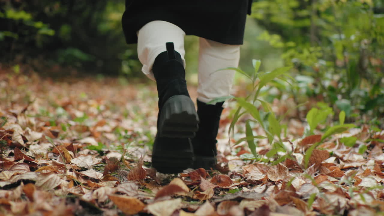바삭거리는 낙엽 카펫이 깔린 숲길을 검은색 부츠를 신은 외로운 소녀가 천천히 거니는 뒷모습, 느긋하고 아늑한 산책, 클로즈업된 발걸음, 가을 분위기, 평온한 자연 속으로의 탈출, 부드러운 보케 지면 시점
