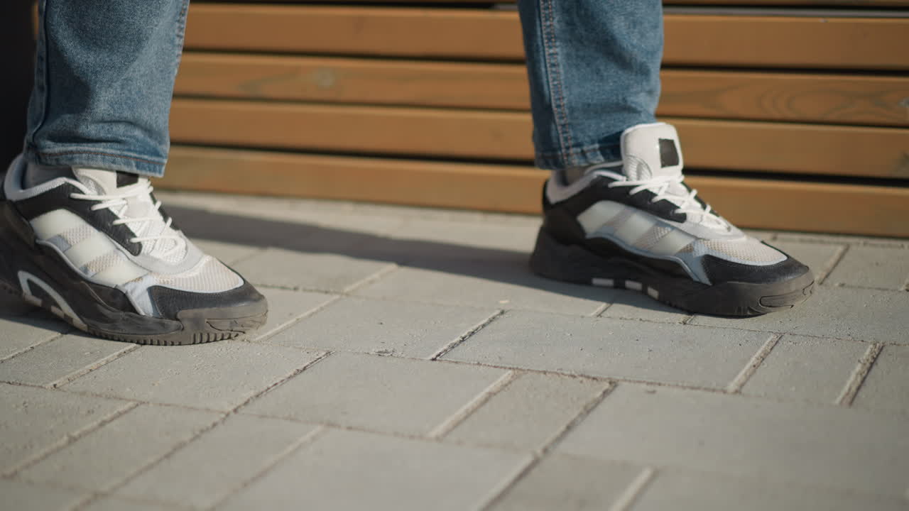 close up of person in jeans and black white canvas walking toward wooden bench to sit while afternoon sun casts strong shadows on paved ground in peaceful modern outdoor environment