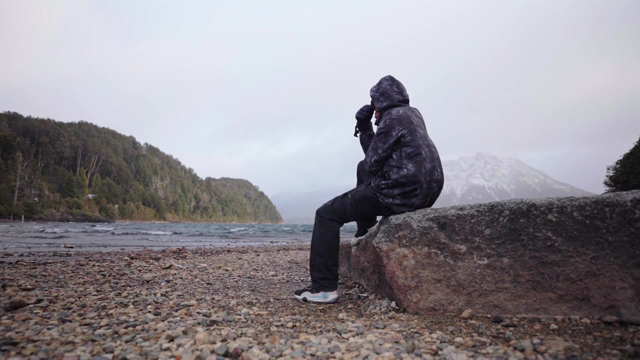 Argentina Patagonia. Uomo in giacca invernale seduto da solo su un masso sulla riva ventosa del Lago Nahuel Huapi, contemplando la natura.