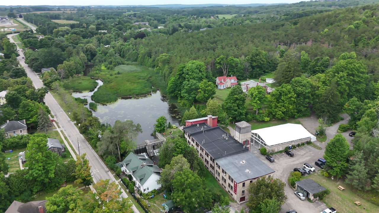 Drone orbiting shot over The Alton Mill complex in Caledon showing historic buildings, road access, small pond and surrounding forest landscape.