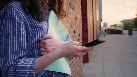 estudiante caucásica de sexo femenino irreconocible de pie con un libro y navegando por el teléfono móvil