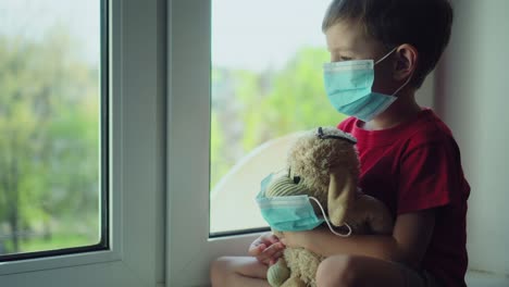 niño y su oso de peluche ambos en máscaras médicas de protección se sienta en el alféizar de la ventana y mira por la ventana 01