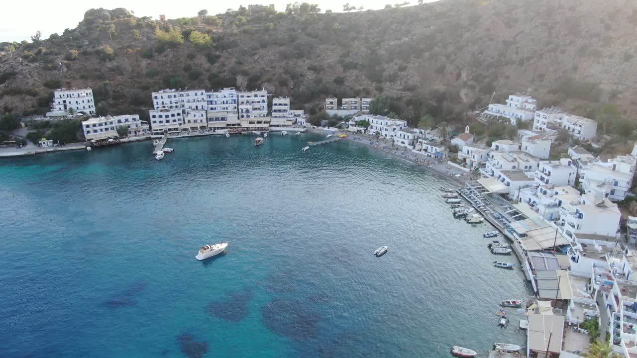 Drone view in Greece panning up over clear blue sea in Loutro small white house town and small boats next to a hill on a sunny day