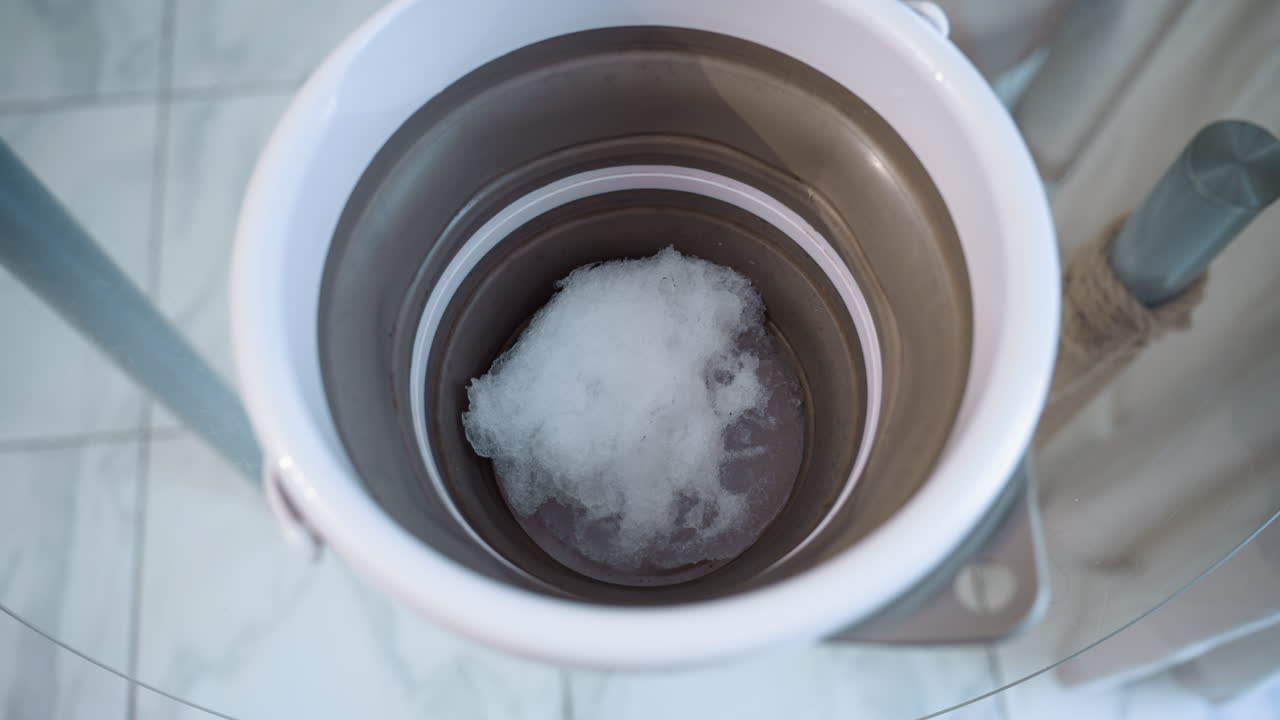 High angle shot showing white and brown bucket containing melting snow placed on plastic table under soft light. Blurred background highlights texture and water droplets inside vessel