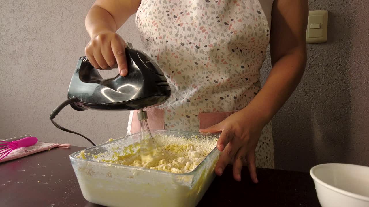 Latinamerikansk kvinna iklädd förkläde förbereder en kaka, blandar alla ingredienser i en behållare med mixern