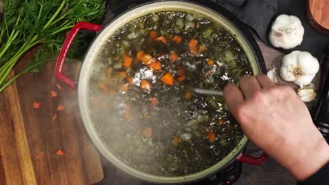 cooking italian wedding soup and adding in the green spinach to the red stock pot. spinach being added into a simmering pot. using a wooden spoon to mix the soup while its simmering.