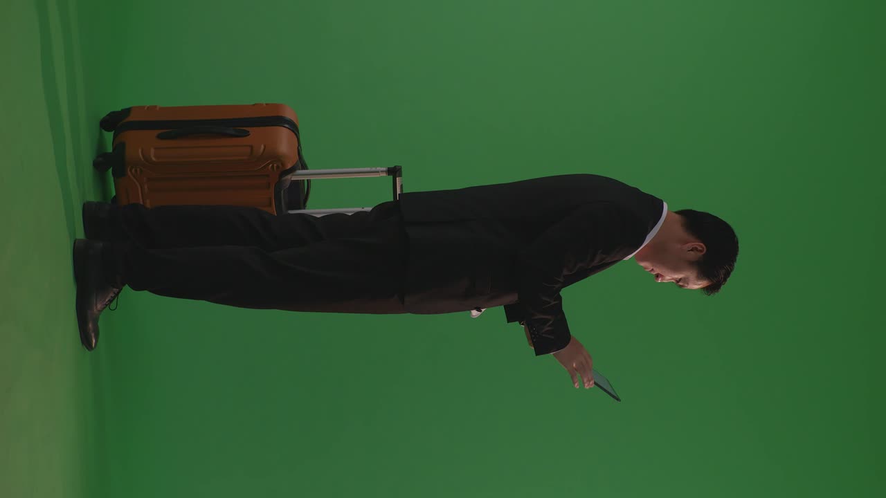Full Body Of Asian Businessman Traveler With Luggage Using A Tablet While Standing In The Green Screen Background Studio, During Sunset Time