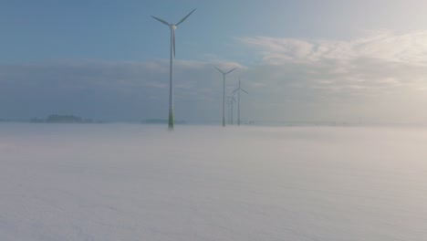 aerial view of wind turbines generating renewable energy in the wind farm, snow filled countryside landscape with fog, sunny winter evening with golden hour light, wide drone shot moving forward low