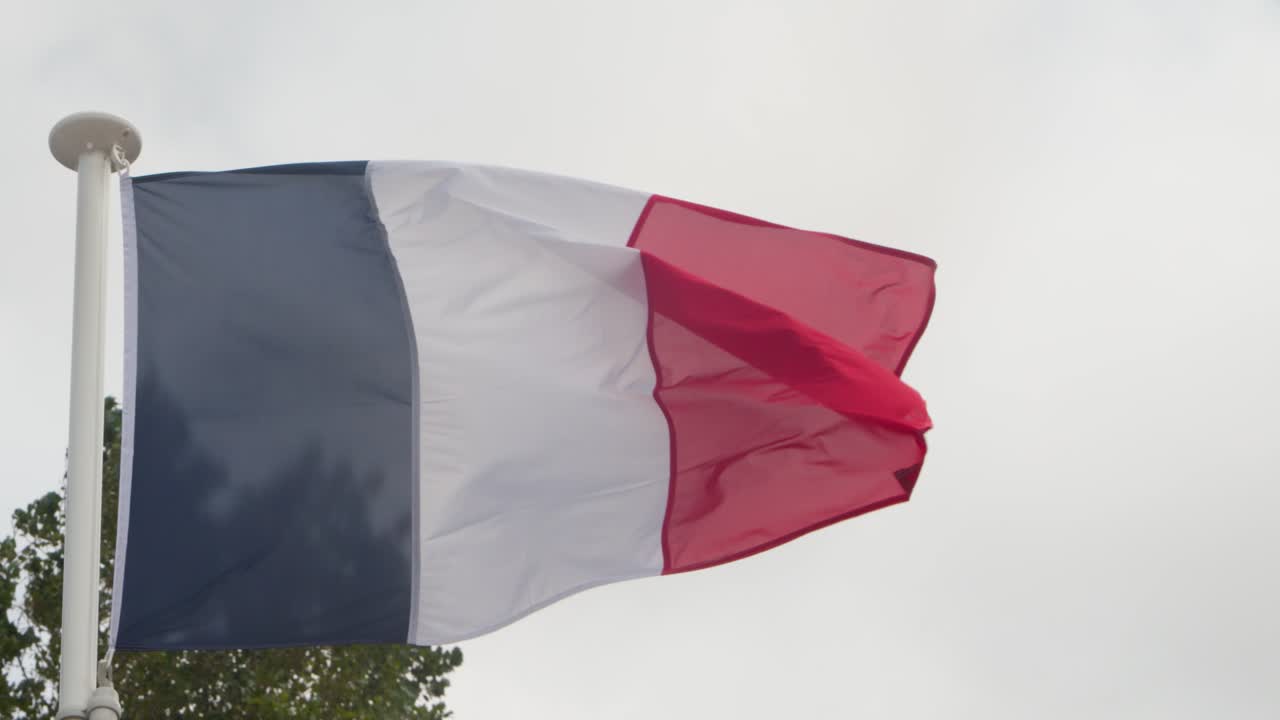 Bandera francesa ondeando en un mástil con fuerte viento, cielo nublado, área conmemorativa al aire libre, toma estable