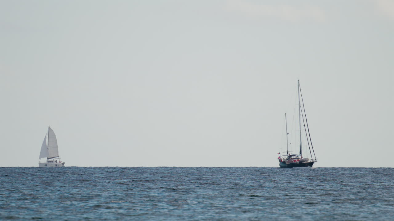 Wide shot of two sailboats navigating the calm blue sea under a pale sky