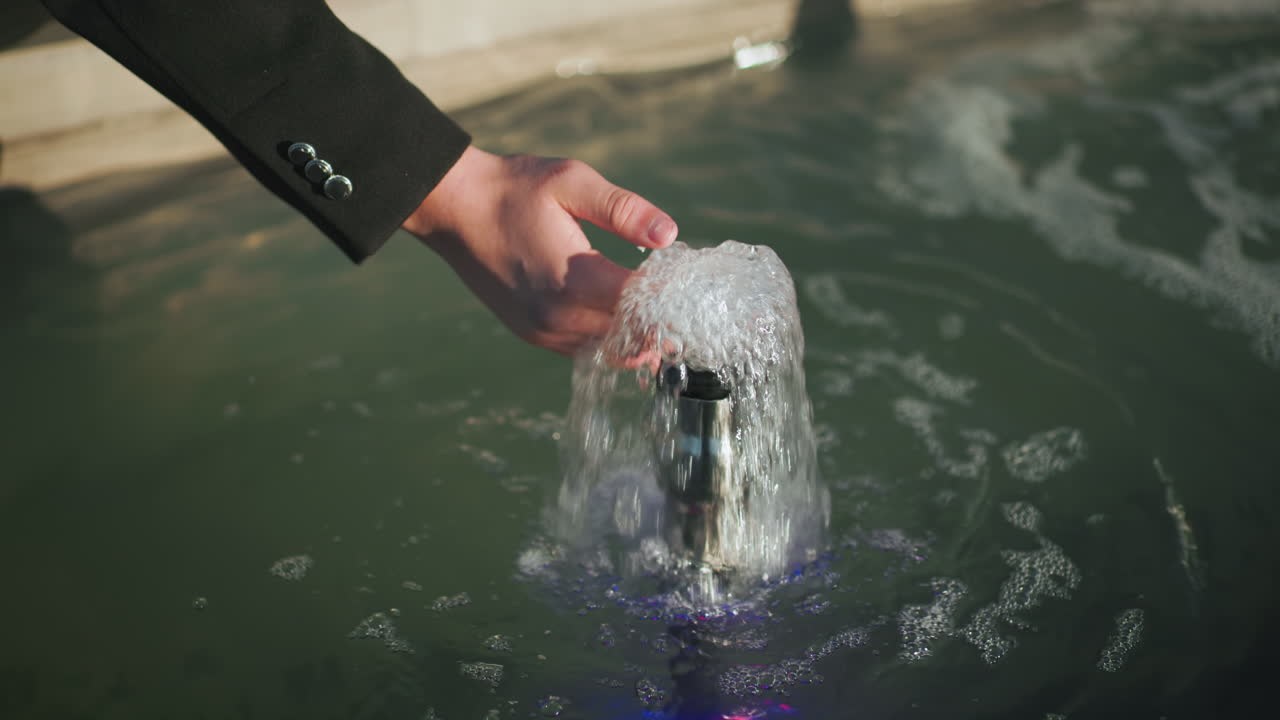 검은 코트를 입은 남자의 손이 작은 분수에서 흐르는 물을 만지고, 걱거리는 물 표면과 반사에 대한 상세한 시각