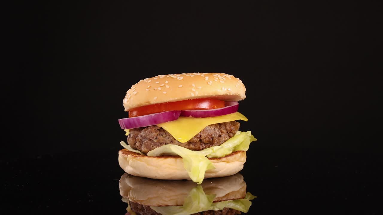 A juicy beef cheeseburger with lettuce, tomato, onion, and cheese is slowly zoomed in on, under bright studio lighting and a black backdrop