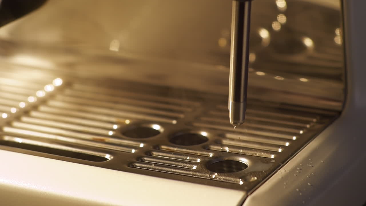 Inquadratura ravvicinata al rallentatore di una lancia vapore di una macchina da caffè espresso che rilascia vapore caldo sopra la vaschetta raccogligocce metallica. Riflessi di luce calda e vapore che sale evidenziano il flusso di lavoro del barista e la preparazione della schiumatura del latte.