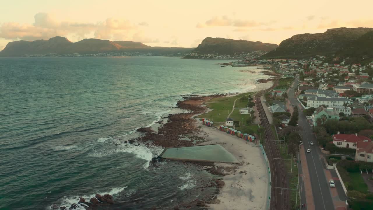 Aerial view of coastal town at sunset with road along shoreline, houses on hillside, rocky beach, tidal pool, calm sea, and pastel sky with scattered clouds.
