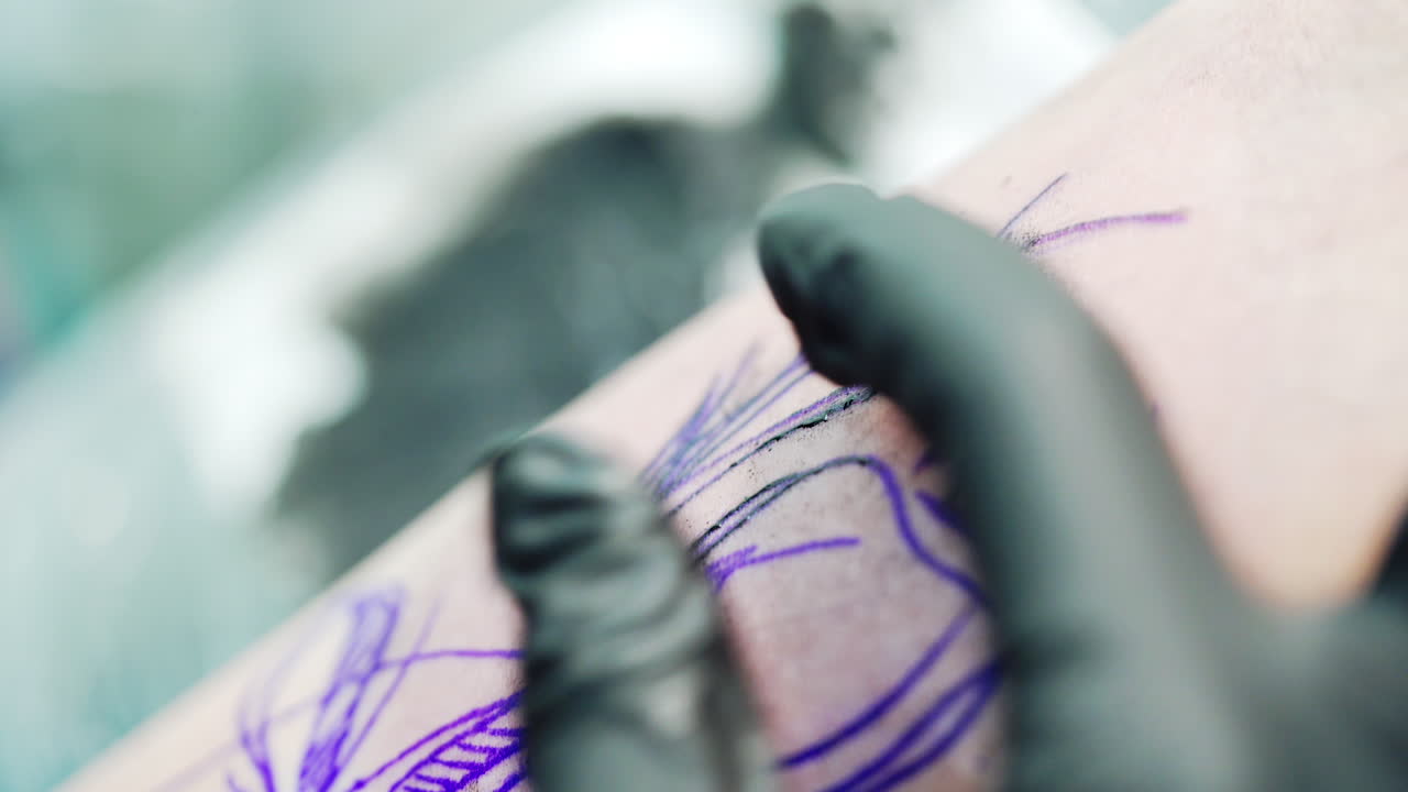 Blue sketched image on skin is covered with black ink from tattoo machine. Gloved hands of tattoo artist stretch the skin white drawing. Close up.
