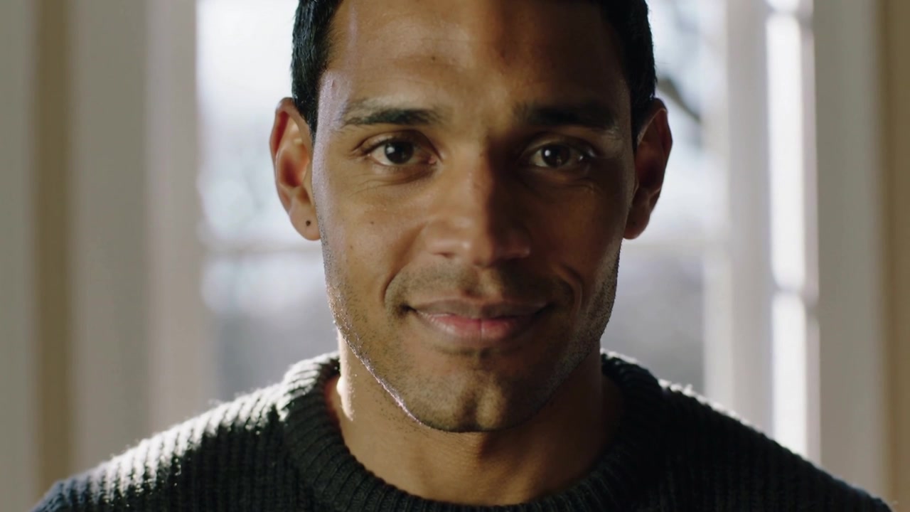 Moving camera creating intimate close-up, man in sweater holding eye contact and smiling at window