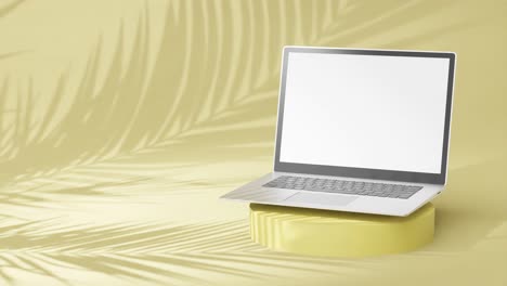 portátil plateado elegante en un pedestal redondo, pantalla vacía con sombras de palma sobre fondo amarillo