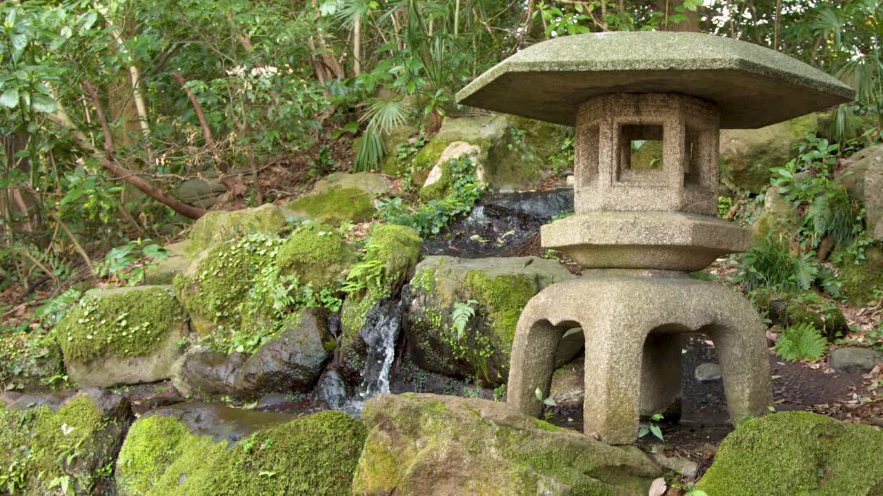 Inquadratura media fissa di una lanterna di pietra accanto a una cascata in un lussureggiante giardino giapponese.