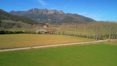 Drone-tracking-from-the-air-of-a-cyclist-on-a-narrow-road-in-an-idyllic-landscape,-green-cultivated-fields-and-mountains-in-the-background-in-La-Garrotxa