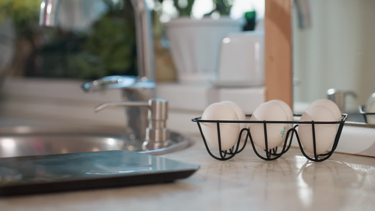 Close up of silver kitchen tap beside soap dispenser with neatly arranged white eggs in black wire rack on countertop, blurred background showing potted plant and window light