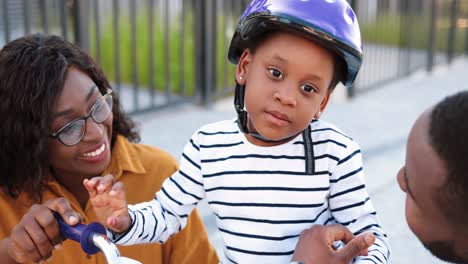 close up of small african american cute girl in helmet sitting on bike and listening to her smiled parents