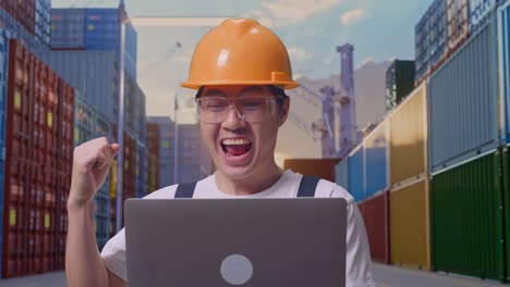 primer plano de hombre asiático trabajador con gafas de protección y casco de seguridad mirando la computadora portátil luego gritando gol celebrando mientras estaba de pie en el depósito de contenedores