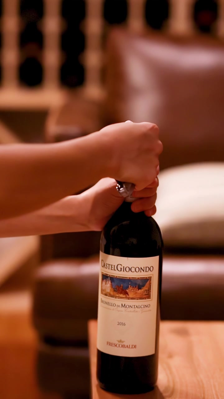 Una persona utiliza un sacacorchos para abrir una botella de vino tinto sobre una mesa de madera en una bodega con iluminación cálida, con un suave desenfoque de fondo.