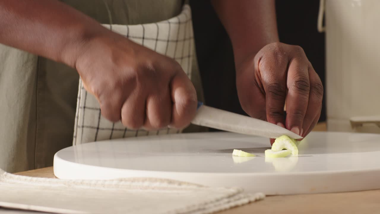 Man wearing checked apron chopping celery at kitchen counter on white cutting board with blue knife