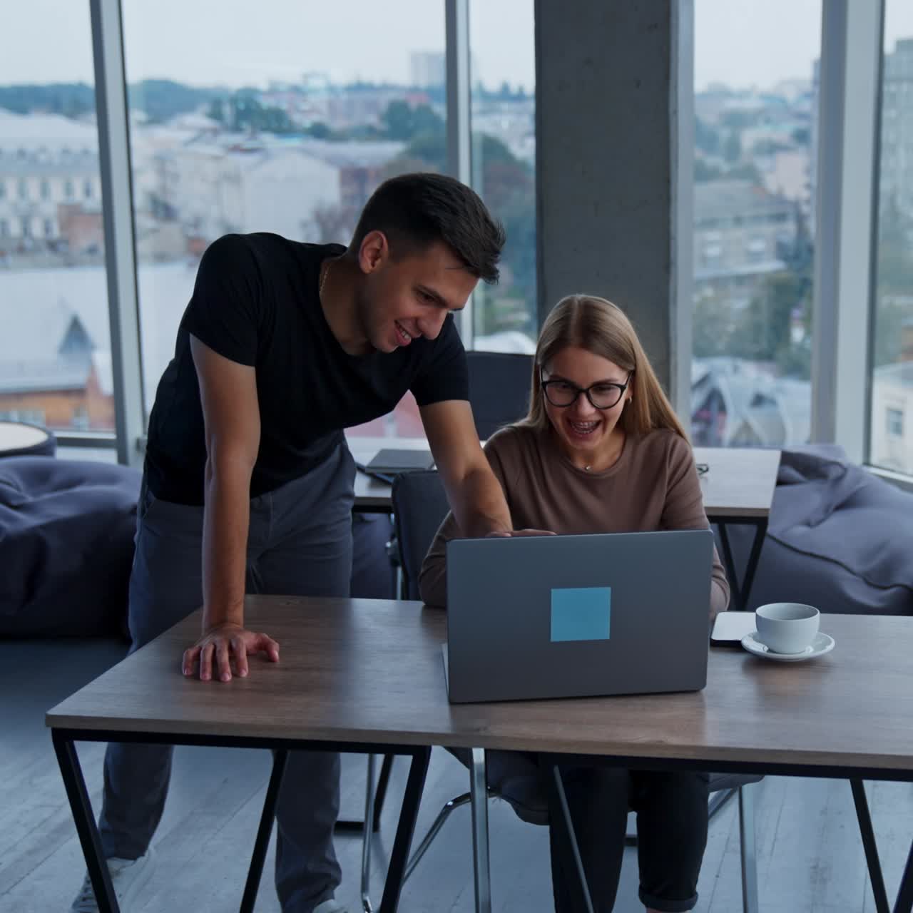 Un collega si china sulla collega seduta alla scrivania. Persone che lavorano insieme discutendo di qualcosa e guardando un portatile sorridendo