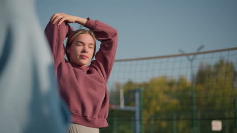 young athlete exercising outdoors with sunlight highlighting her face, a partial view of someone nearby in the background, showcasing an active, energetic atmosphere in a sports arena