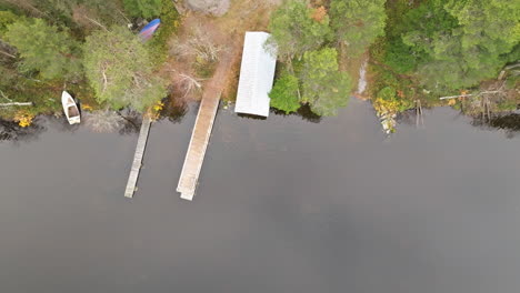 Sweden - Autumn Forest Reflects on the Water by a Lakeside on a Cloudy Day, Complemented by a Floating Wooden Pier and Traditional Fishermen&#039;s Houses - Aerial Sideways