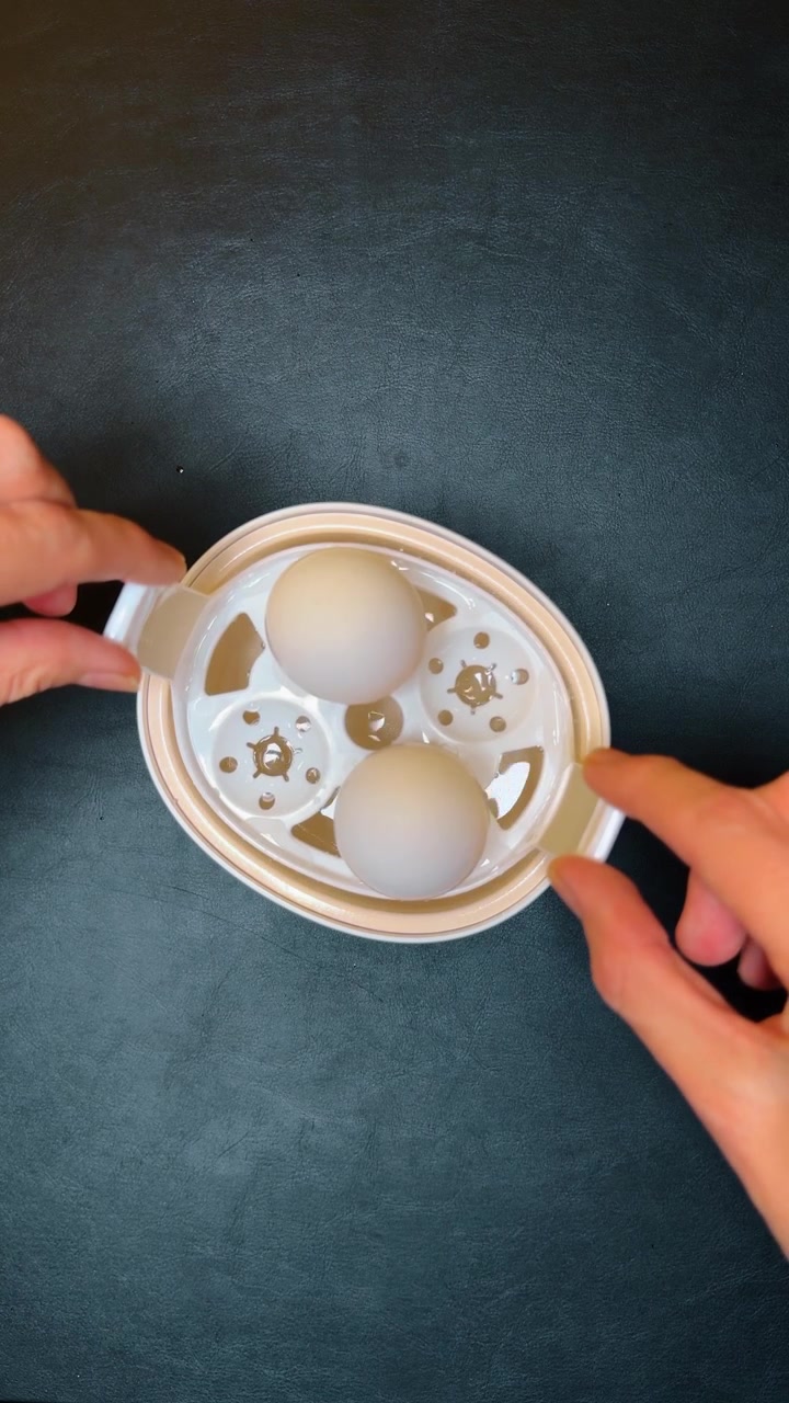 Placing egg tray into electric steamer with water. Top view of human hands placing plastic tray with two eggs into cooker base filled with water.