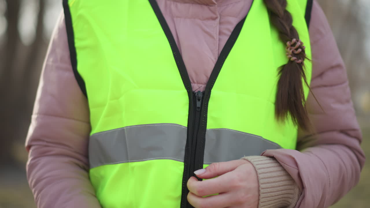 Close-up of woman in pink puffer jacket zipping bright neon yellow reflective safety vest outdoors, preparing for increased visibility and protection during outdoor work or volunteering in cool weather