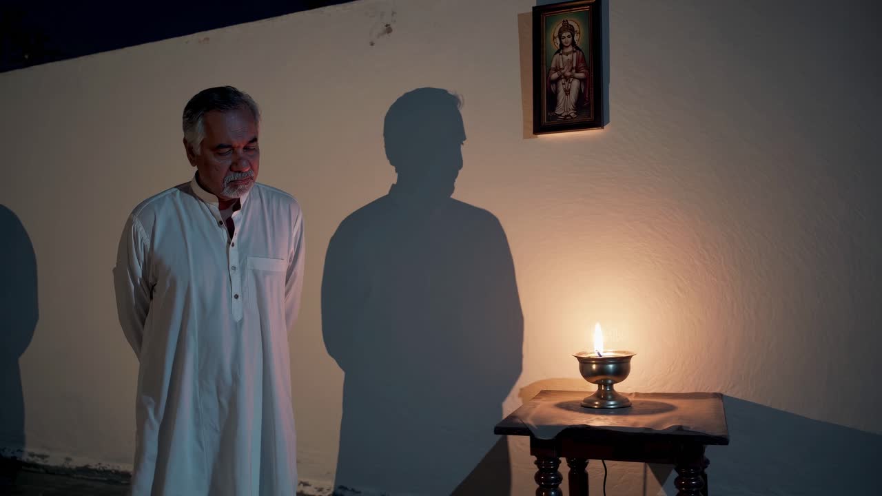 Man in traditional attire stands in contemplation beside a lit oil lamp, casting shadows on the wall, creating a serene atmosphere of reflection and spirituality