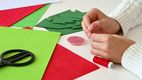 female hands get sewing thread in needle to make christmas crafts with colored fabrics