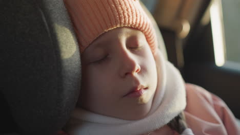 close-up of a young girl peacefully sleeping in a car, wearing a pink knit hat and scarf, with sunlight gently illuminating her face