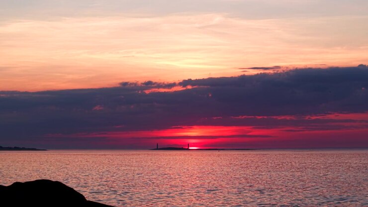 Un'alba vivida illumina gli iconici fari gemelli dell'isola di Thacher al largo di Rockport, MA. L'oceano calmo riflette il cielo infuocato, atmosfera tranquilla, nuvole drammatiche. Paesaggio costiero grandangolare, nessuno visibile.
