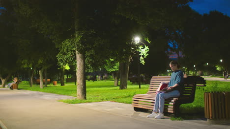 young woman sits outdoors on bench at dawn, opening laptop in serene park setting, background shows trees, a light pole, greenery, and people strolling and riding bicycles