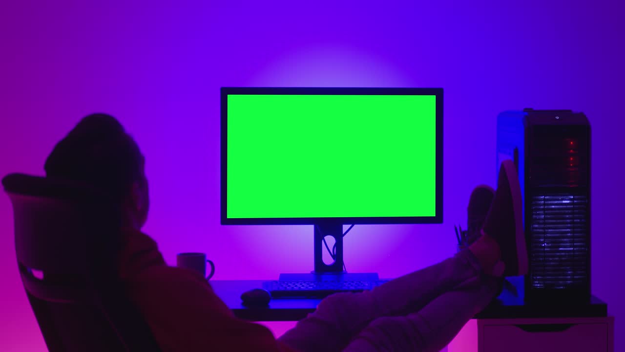 Young man freelancer resting on a chair, looking at a computer monitor with a green screen chroma key, indoors at night in room filled with neon light.t.
