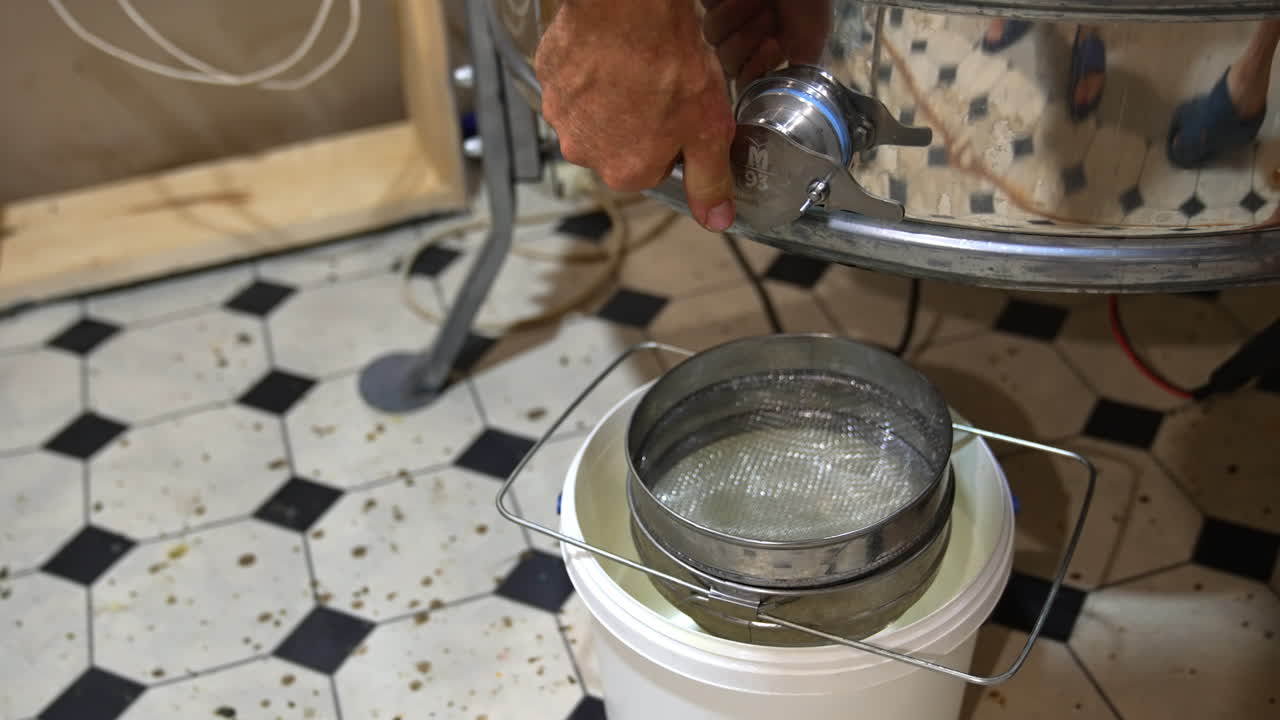 Man opens the faucet at honey extracting machine. Fresh golden organic liquid honey flows into the bucket standing under the equipment.