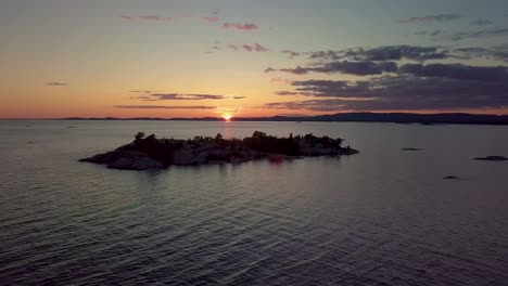 flying away from rocky pine tree island in blue lake at sunset, drone aerial wide dolly out