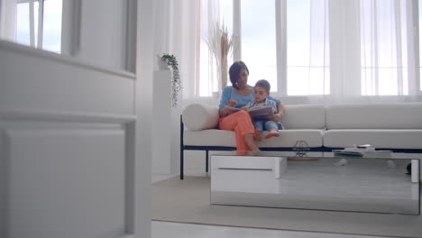 Happy-mother-and-little-son-reading-a-book-in-the-morning-together-in-the-living-room-at-home.-family-activity-concept.