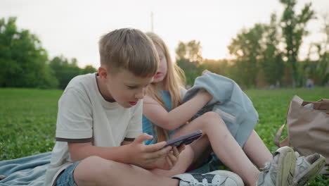 boy and girl seated on grass during park outing, boy holding phone focused on screen while girl lifts arm to put on sweater, casual summer clothing, soft sunset light, playful sibling interaction