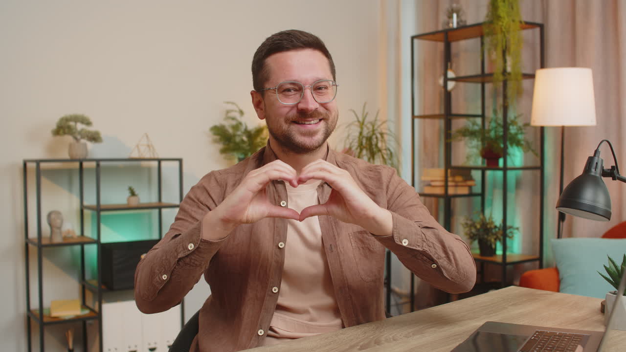 Happy caucasian young man makes symbol of love showing heart sign to camera at home office desk